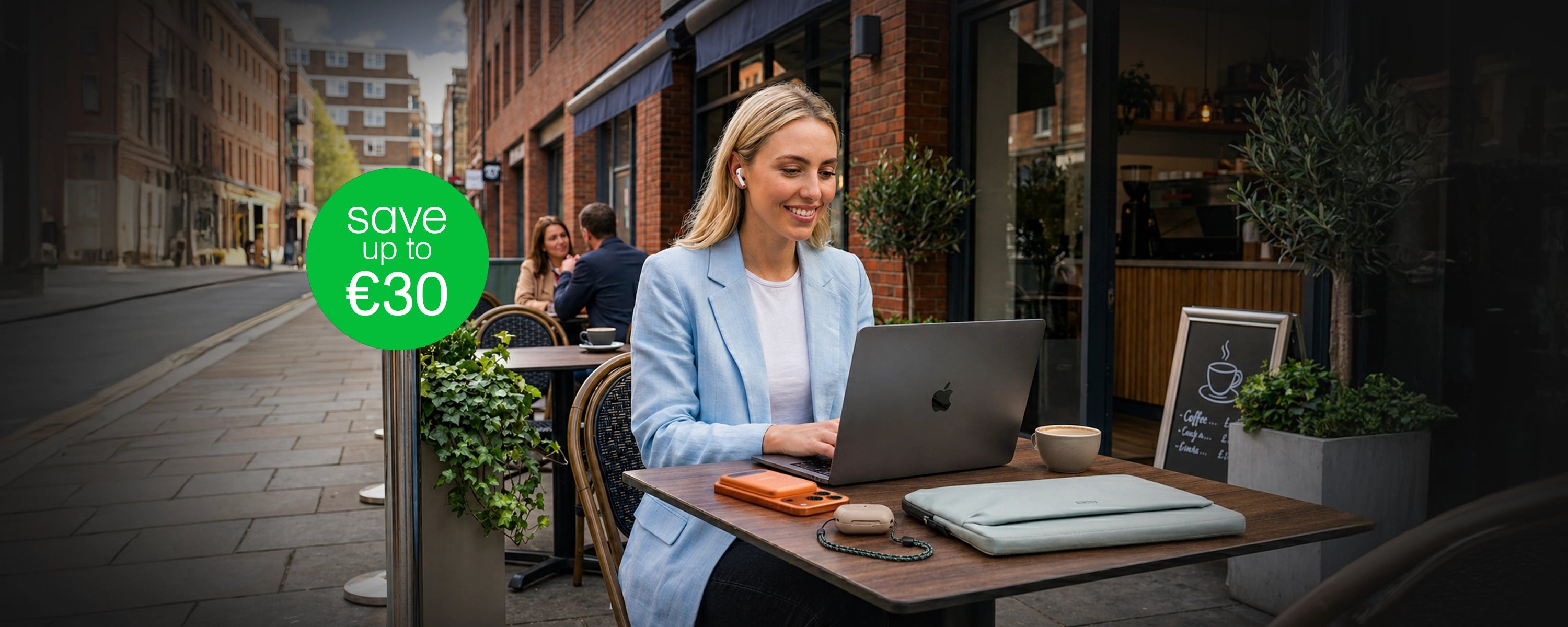 Woman using a laptop at an outdoor cafe with a promotional sign for savings.